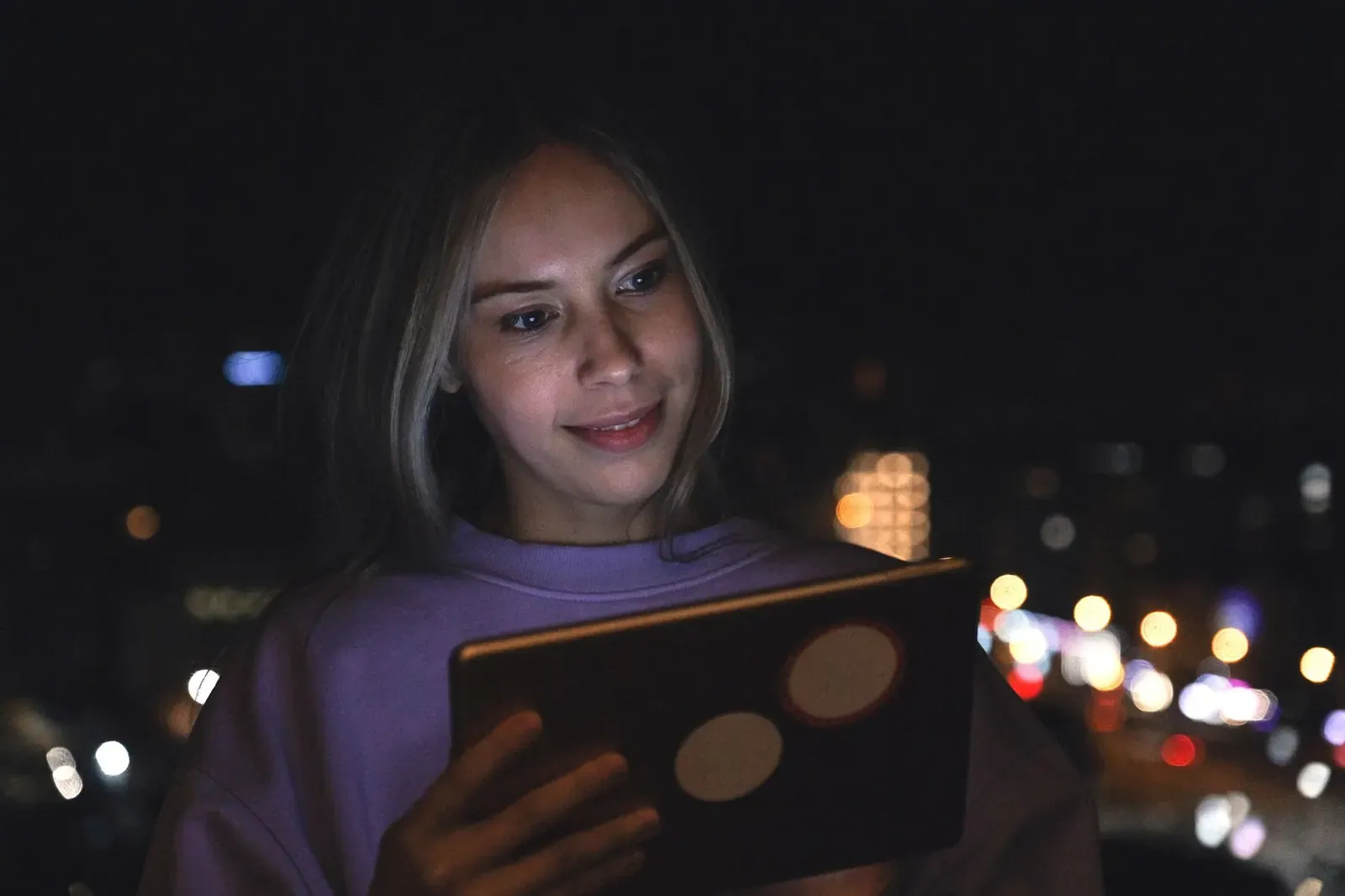 Une femme regarde une tablette le soir, des lumières de ville floues derrière elle