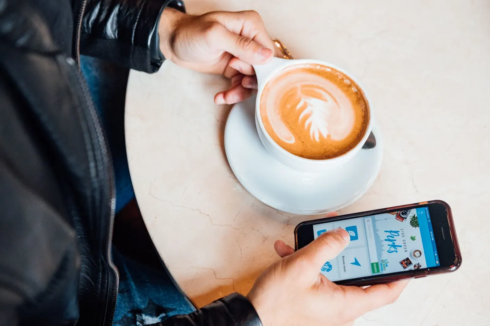 Une main tient un smartphone à côté d'un café au lait, sur une table de terrasse