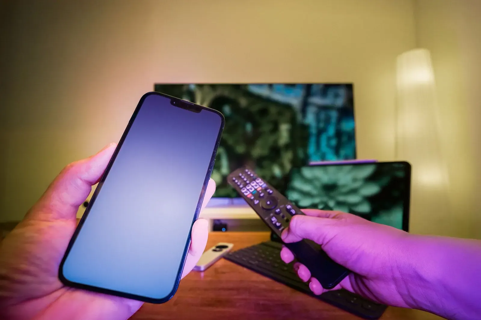 Une main tient un smartphone et une télécommande devant un téléviseur allumé